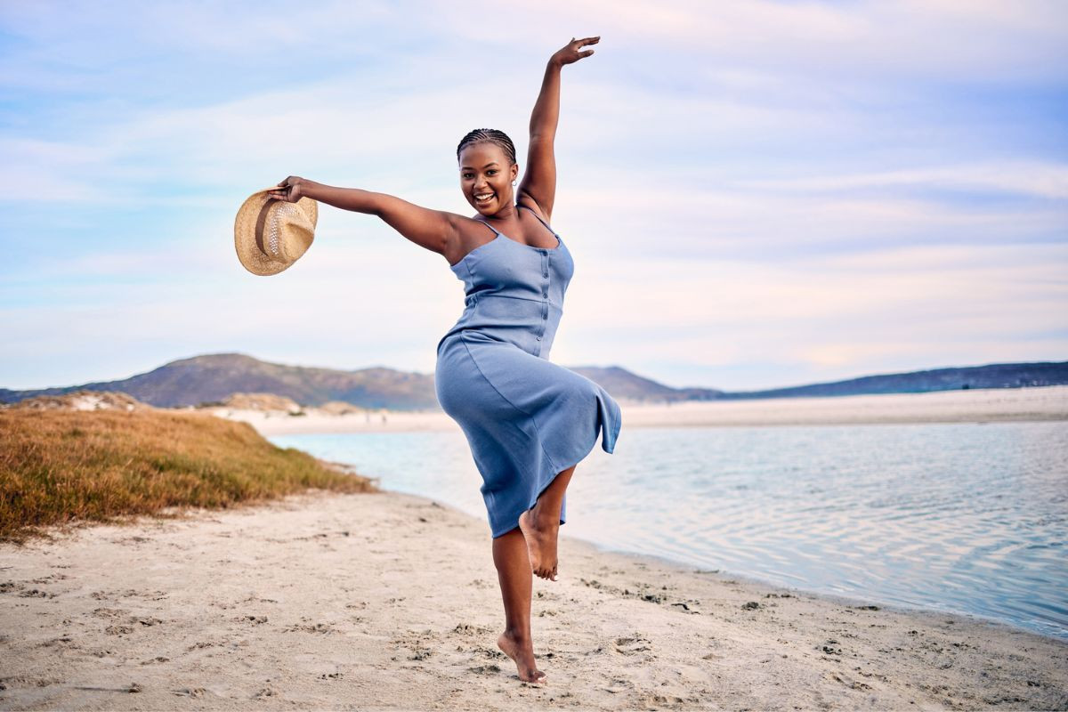 african amercain woman dancing on a beach in sundress and hat