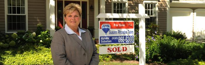 Susann Rhoades standing by for sale sign Logo